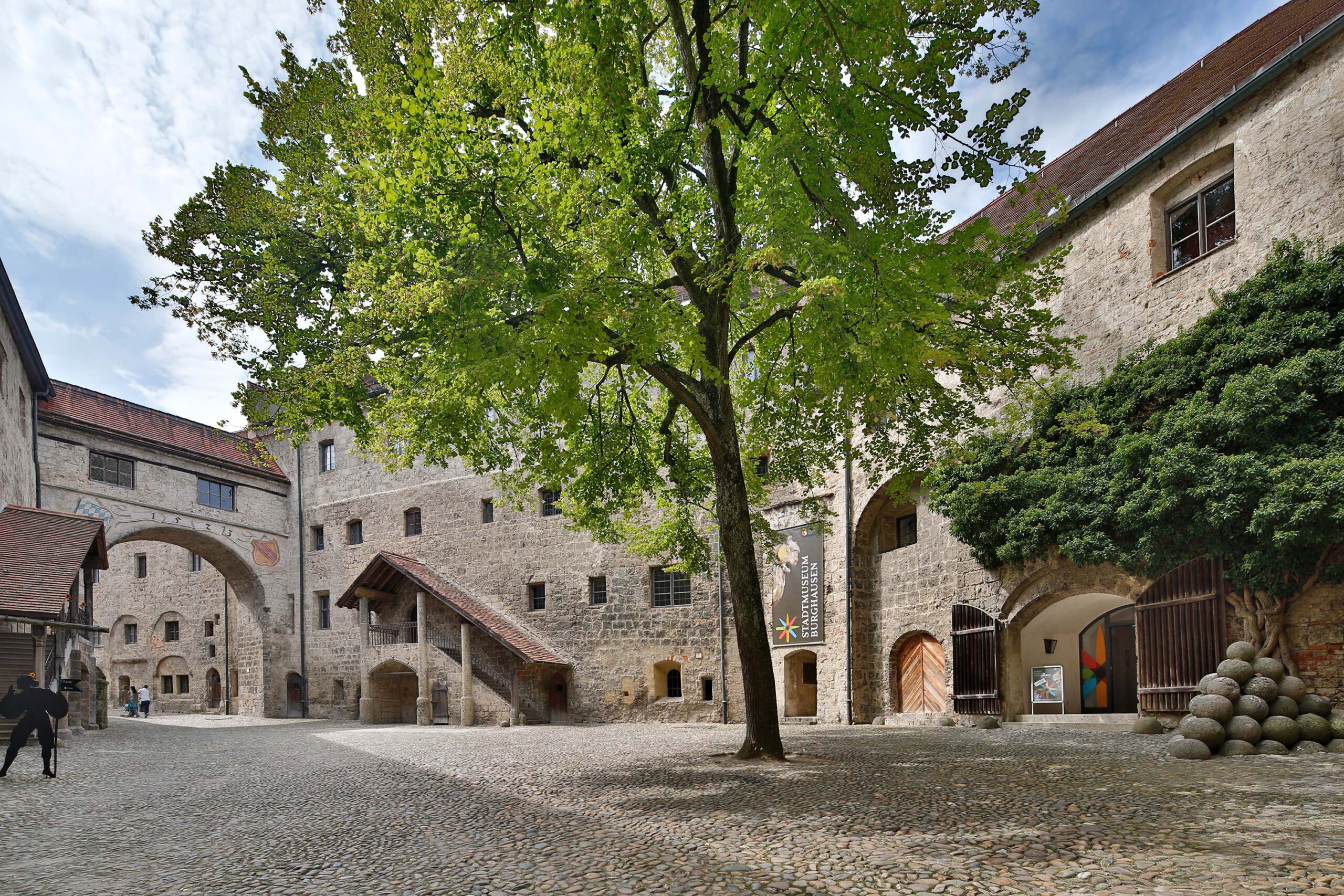 Innenhof einer Burg aus Stein. In der Mitte des Hofs steht ein Baum. Rechts ist ein Eingang mit einem Plakat Stadtmuseum Burghausen.